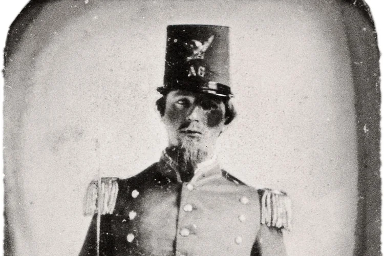 Hazy 1860s portrait of a man in a tall uniform hat marked AG, covered in dust specks