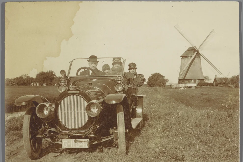 Yellowed 1920s photo of people in an old car near a windmill, with a large water stain in the sky